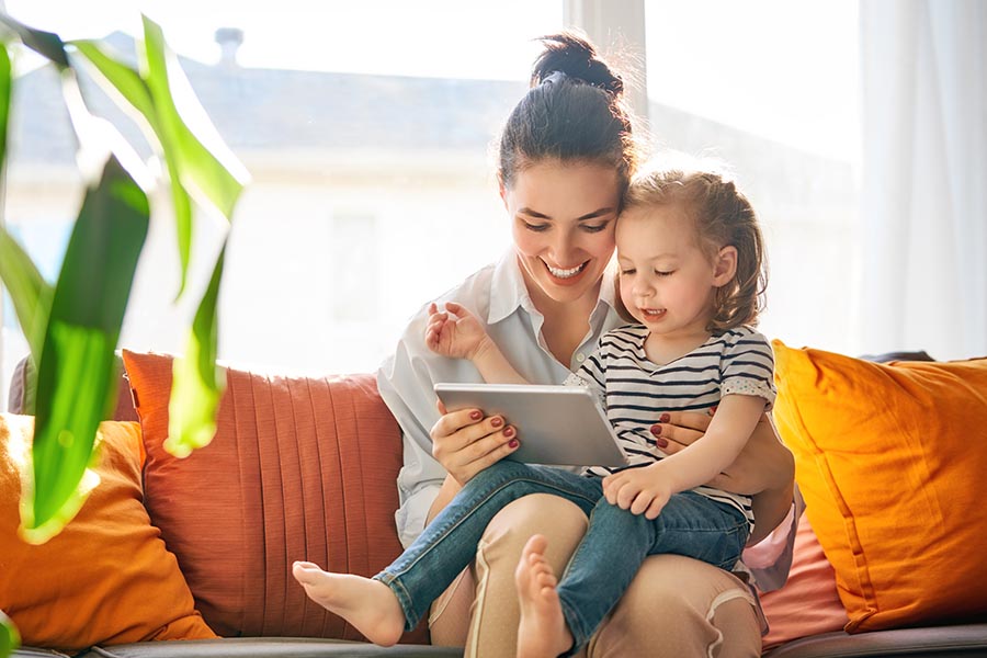 Blog - Mom and Young Daughter Use a Tablet on Their Bright Orange Sofa, a Houseplant in the Foreground